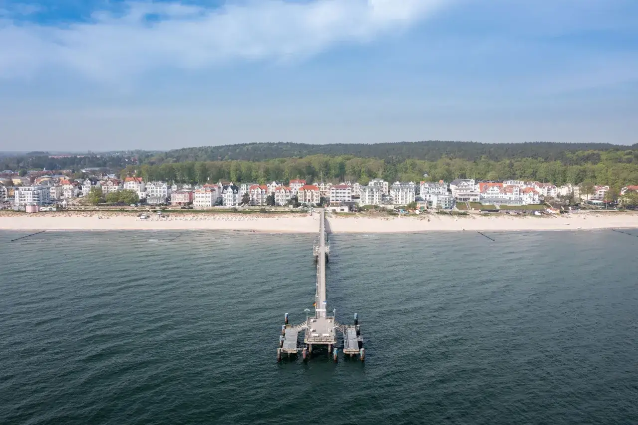 Blick auf den Strand von Bansin mit Seebr&uuml;cke, wei&szlig;en H&auml;usern und gr&uuml;nen W&auml;ldern im Hintergrund.