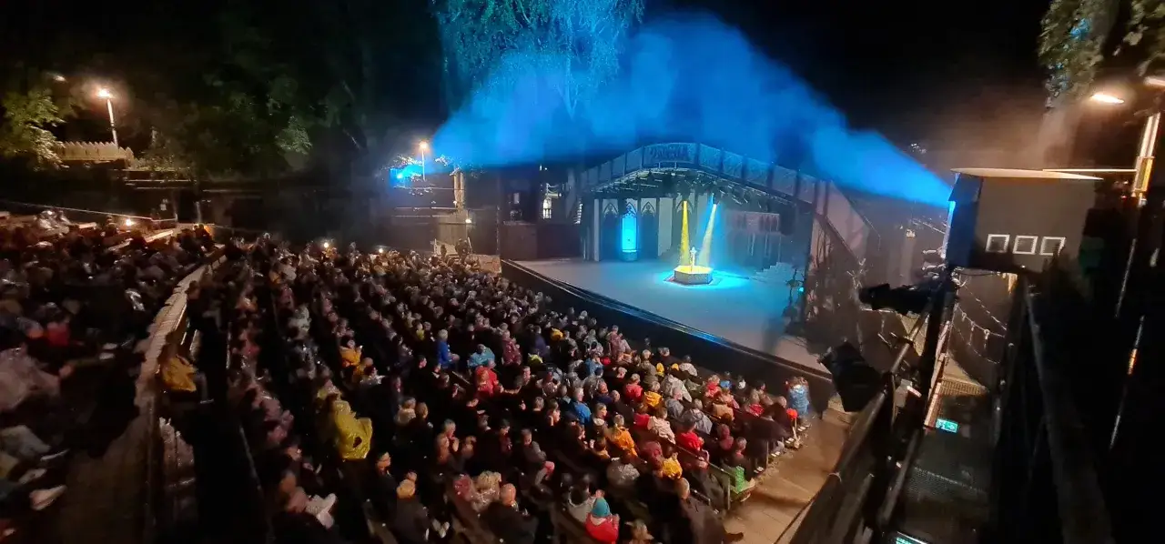 Publikum sitzt gespannt bei den Vineta Festspielen Zinnowitz. Blaue und gelbe Scheinwerfer beleuchten die Bühne, auf der ein Darsteller steht.