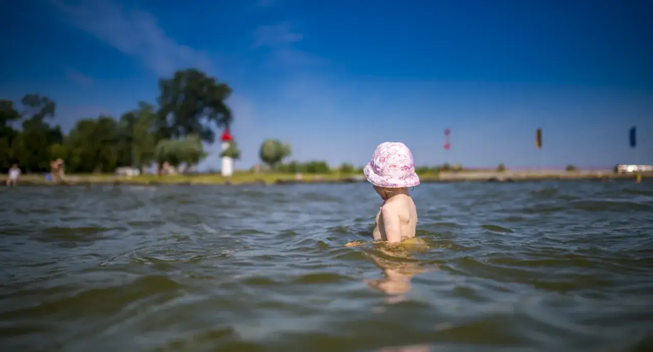 Kleines Kind mit rosa Hut planscht im Wasser am Stettiner Haff Strand. Im Hintergrund B&auml;ume und ein Leuchtturm.