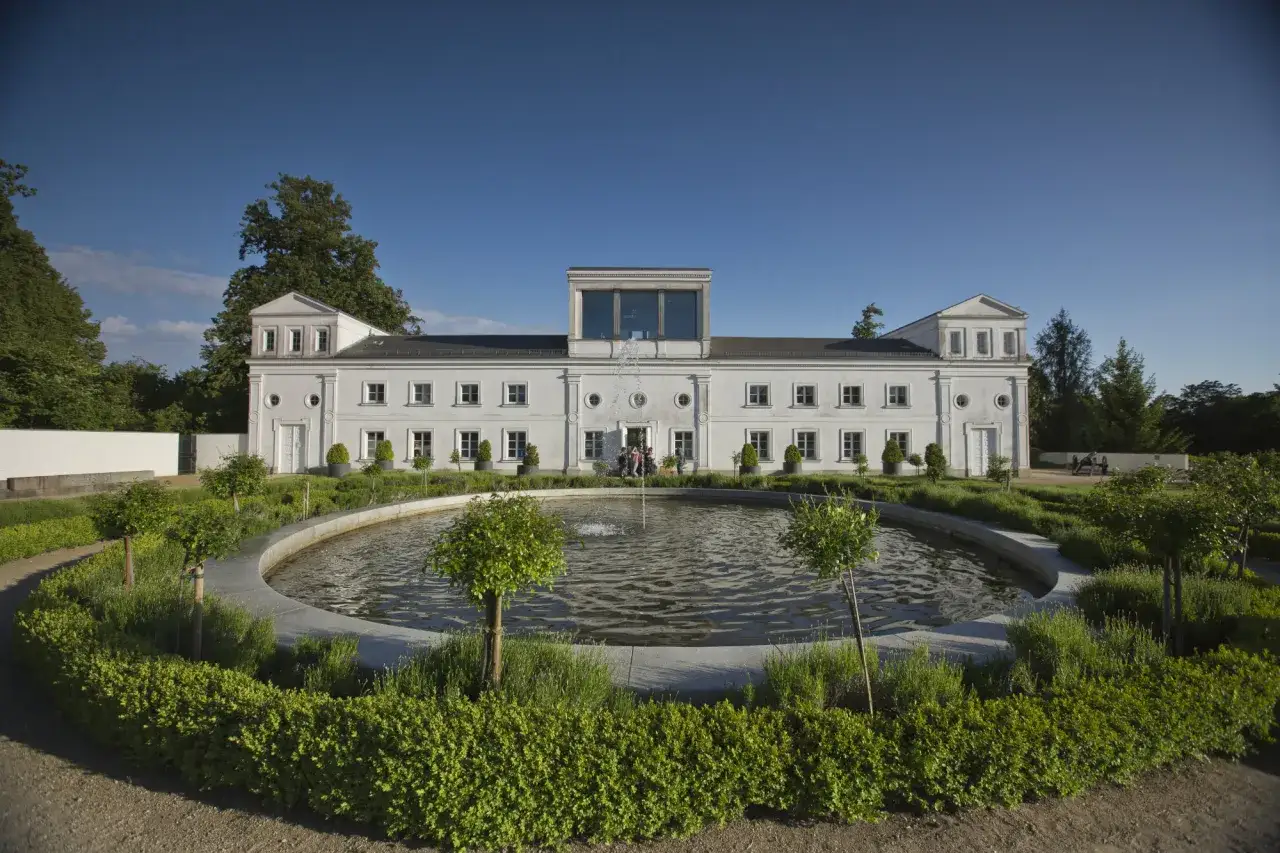Die Orangerie Putbus mit ihrem runden Brunnen und gepflegten Gartenanlagen, ein architektonisches Juwel unter blauem Himmel.