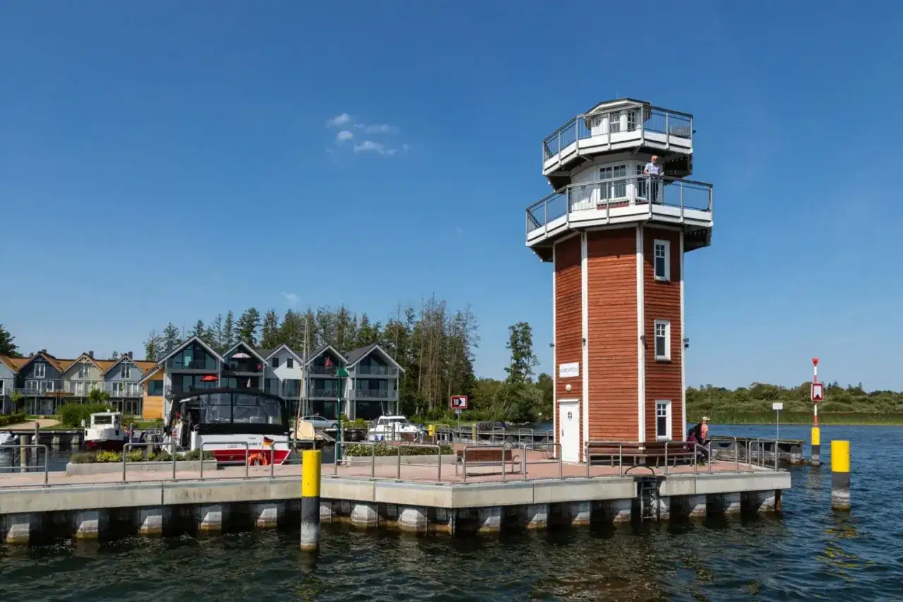 Ein Mann steht auf dem Aussichtsdeck des roten Leuchtturms. Im Hintergrund reihen sich moderne H&auml;user am Wasser. Ein sch&ouml;ner Tag am See mit Promenade.