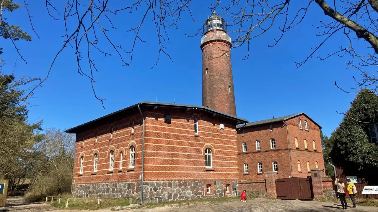 Der Leuchtturm Prerow ragt imposant in den blauen Himmel. Ziegelbauten mit Rundbogenfenstern s&auml;umen seinen Fu&szlig;.