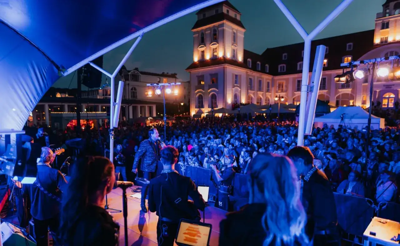 Ein Konzert auf dem Kurplatz in Binz. Ein Sänger steht auf der Bühne, umgeben von einer jubelnden Menge. Die Gebäude im Hintergrund sind beleuchtet.