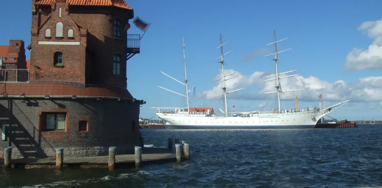Das Segelschulschiff Gorch Fock in Stralsund, ein imposantes wei&szlig;es Schiff mit drei Masten, liegt vor einem Backsteinturm im Hafen.