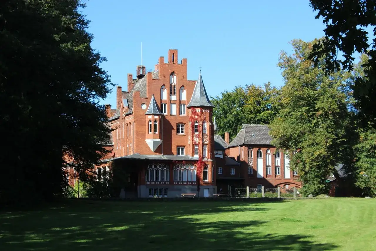 Das Schloss Kalkhorst, ein roter Backsteinbau mit T&uuml;rmchen und Erkern, steht auf einer gr&uuml;nen Wiese, umgeben von B&auml;umen.