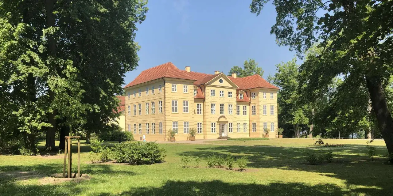 Das Schlossinsel Mirow, ein gelbes Barockschloss mit roten D&auml;chern, steht auf einer gr&uuml;nen Insel, umgeben von B&auml;umen.