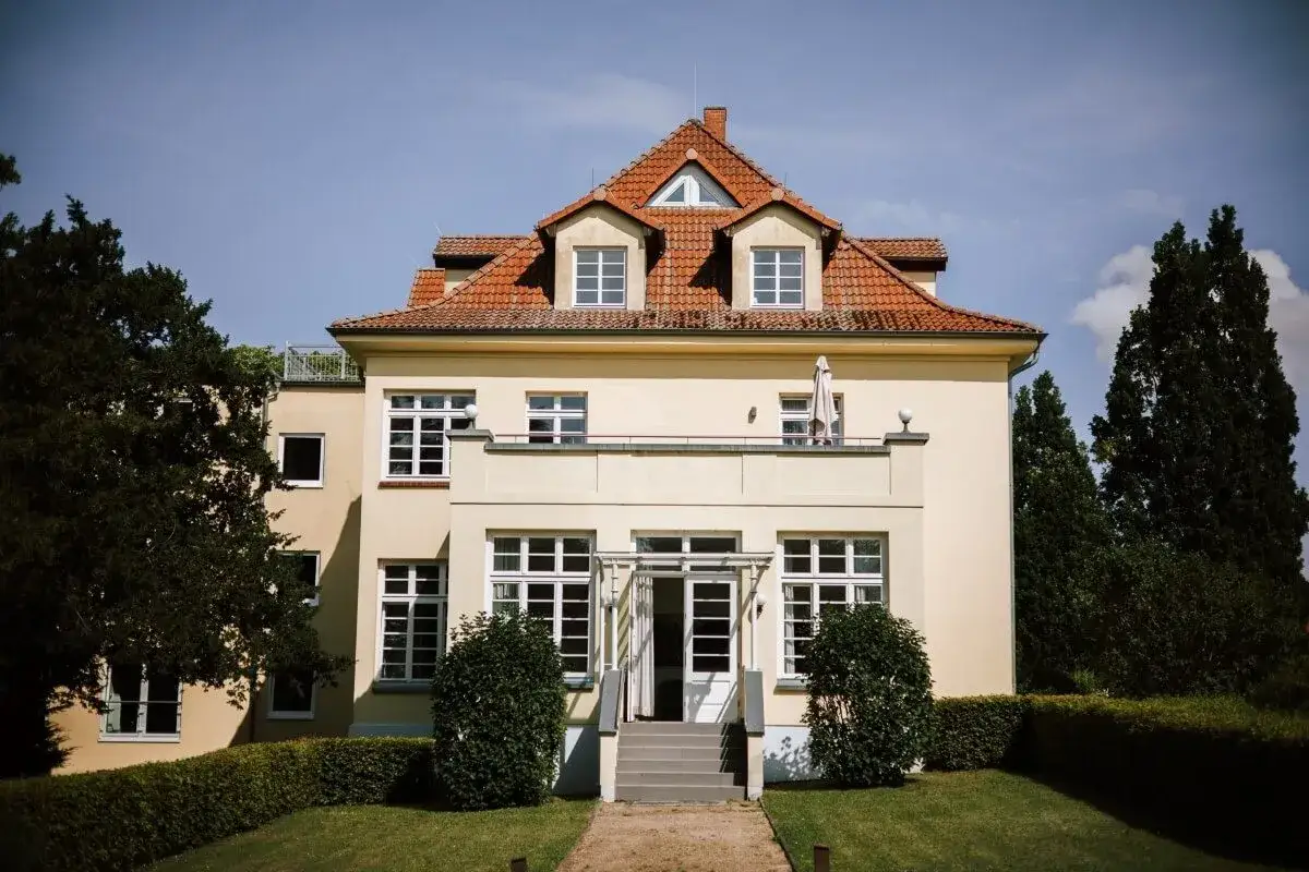 Ein gelbes Haus mit roten Ziegeldach und vielen Fenstern, umgeben von grünen Bäumen und Büschen. Ein gut gremmeliner Ort für einen entspannten Tag.