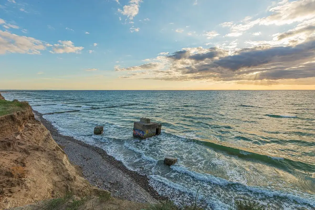 Ein alter Bunker am Strand, umsp&uuml;lt von Wellen. Die Sonne bricht durch Wolken und beleuchtet das Meer.
