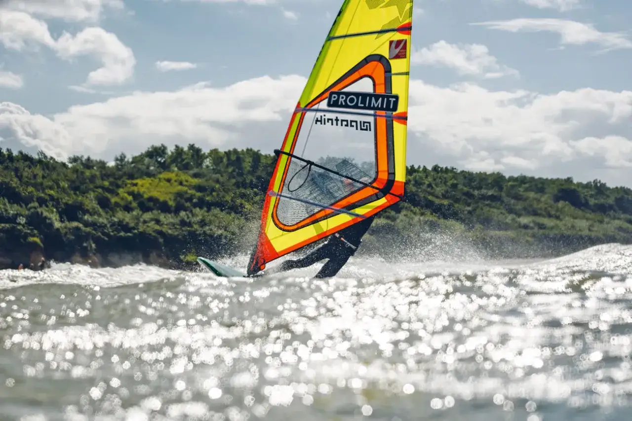 Windsurfer gleitet &uuml;ber die Ostsee, die Sonne funkelt auf den Wellen. Ein bunter Segel dominiert das Bild.
