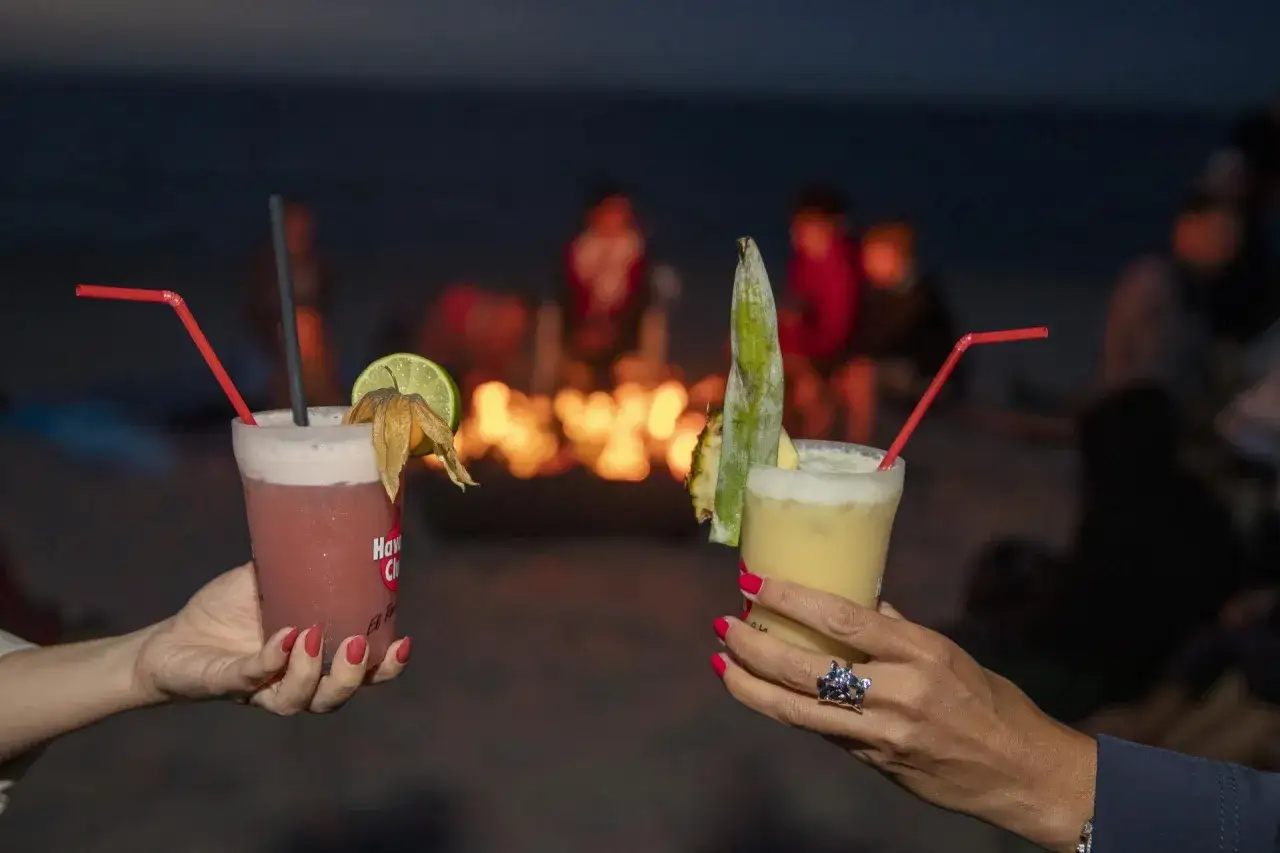Zwei Cocktails am Strand bei Nacht. Im Hintergrund ein Lagerfeuer und verschwommene Silhouetten von Menschen. Perfekt f&uuml;r eine Ostsee Party.