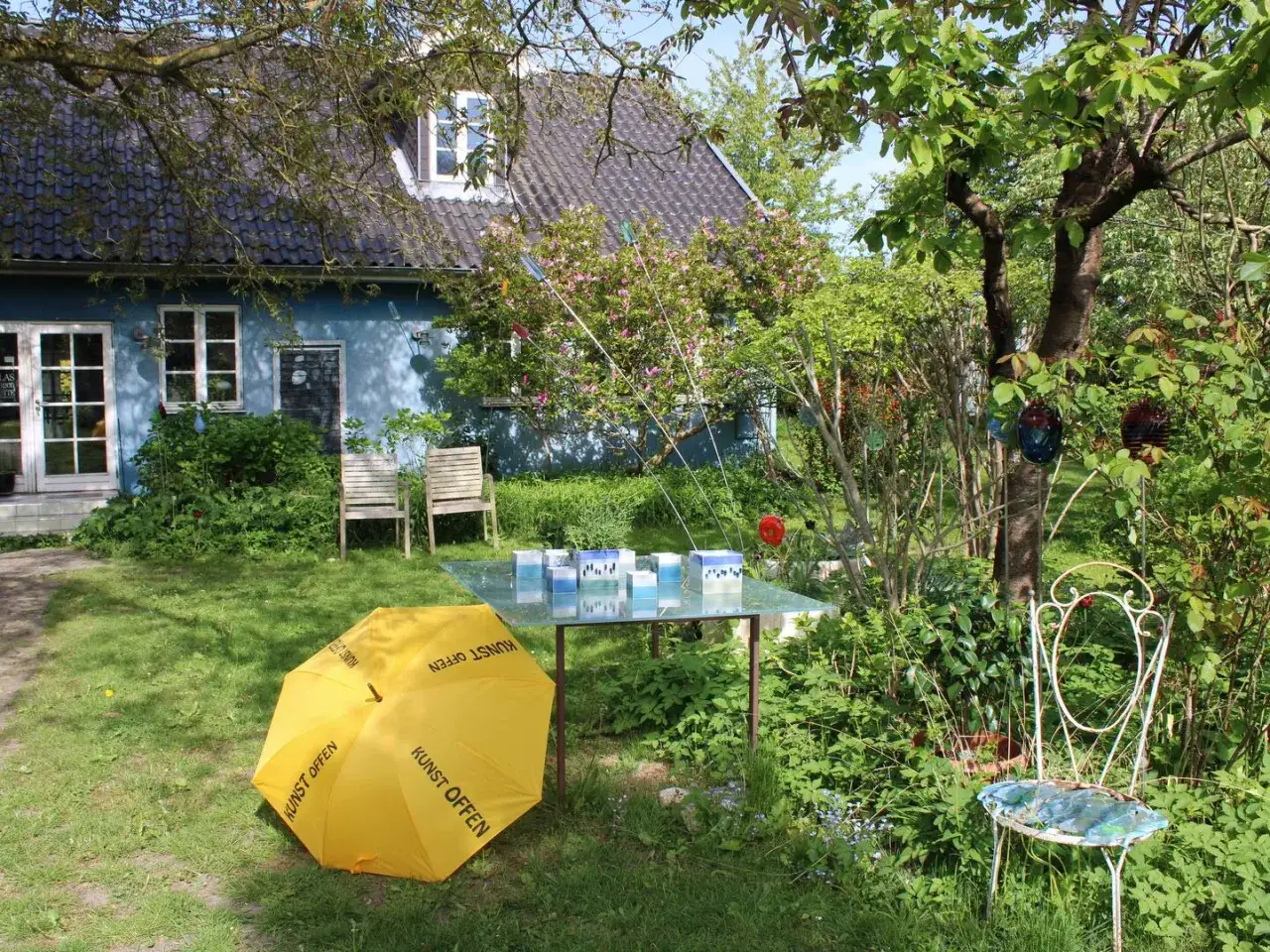 Ein Garten mit blauem Haus, gelbem Regenschirm (Kunst Offen) und Glaskunst auf einem Tisch. Pfingsten 2026 MV.