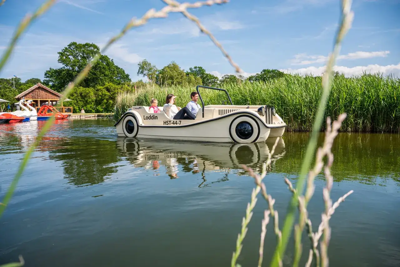 Familie genie&szlig;t eine Fahrt mit einem Auto-Boot von Kikis Bootsverleih auf einem ruhigen See, umgeben von gr&uuml;ner Natur.