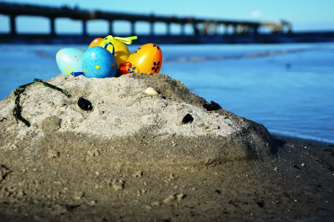 Bunte Ostereier auf einer Sandd&uuml;ne am Meer. Ein Pier im Hintergrund. Perfekt f&uuml;r Veranstaltungen an Ostern am Strand.