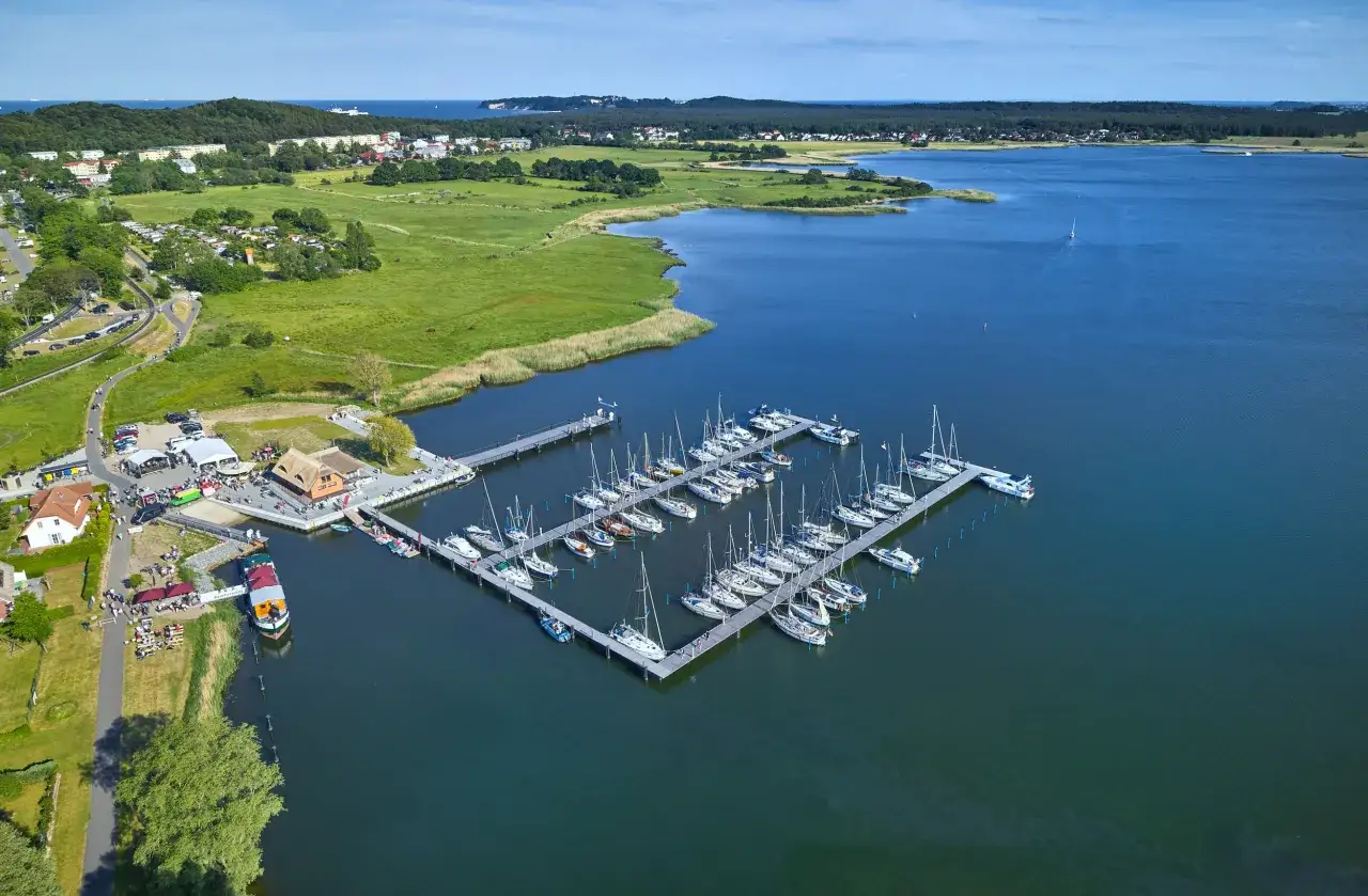 Der Hafen Sellin ist voller Segelboote, die im ruhigen Wasser liegen. Im Hintergrund erstreckt sich eine gr&uuml;ne Landschaft mit H&auml;usern und B&auml;umen.
