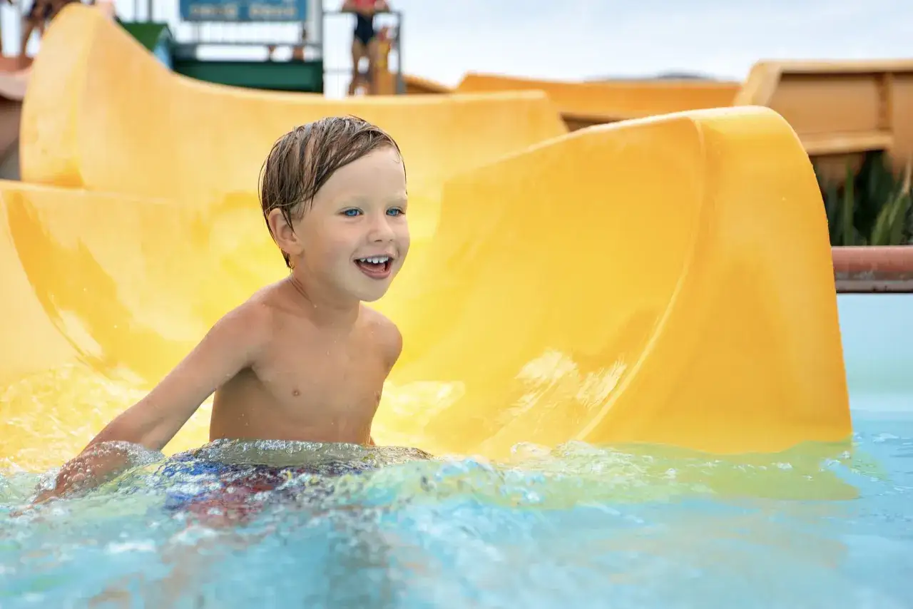 Ein Junge mit blauen Augen lacht fr&ouml;hlich im Wasser am Ende einer gelben Wasserrutsche im Heidebad Torgelow.