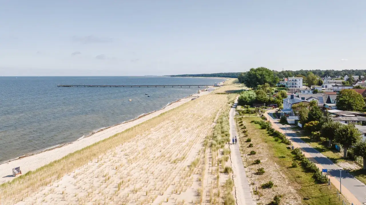 Ein Paar spaziert mit Hund am Strand entlang. Im Hintergrund die Ostsee mit Seebrücke und ein Küstenort.
