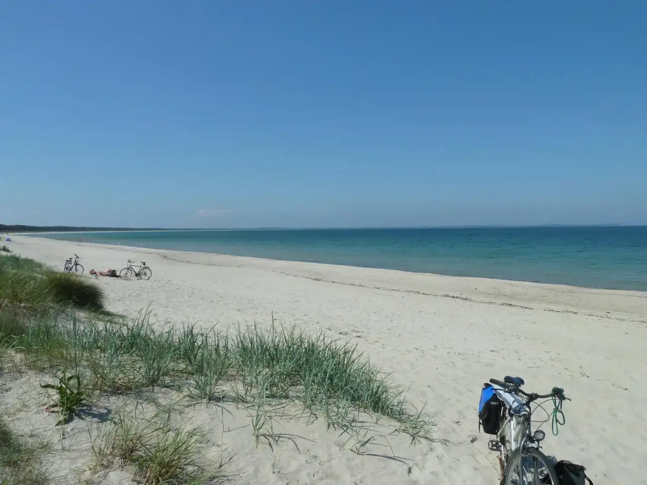 Ein einsamer glowe Strand mit zwei Fahrrädern und zwei Sonnenanbetern. Das Meer glitzert in der Sonne.