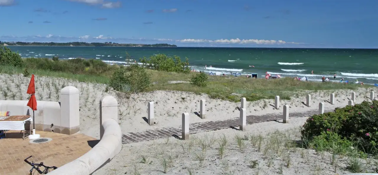 Sonniger Tag am Juliusruh Strand. Menschen genießen das Meer, Strandkörbe und eine Promenade führen zum Wasser.