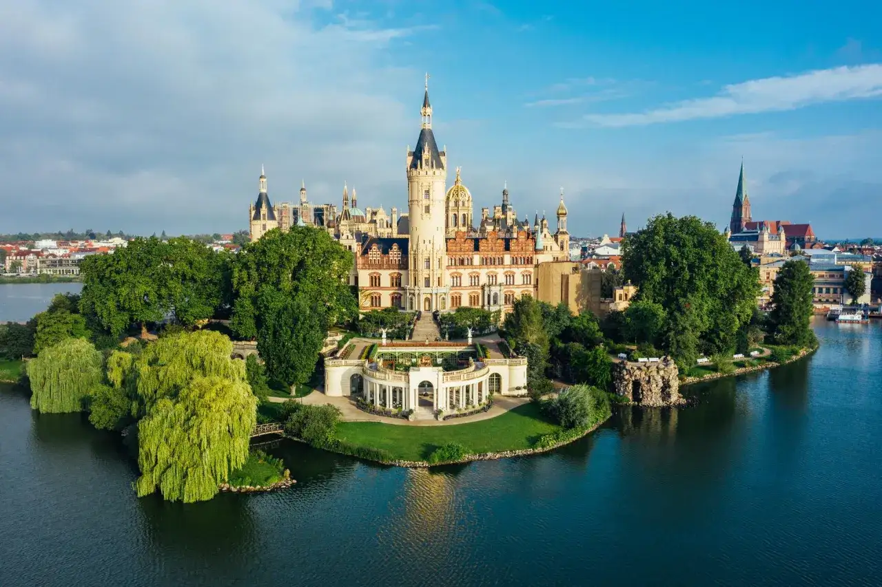 Das Schweriner Schloss, eine Märchenkulisse auf einer Insel, thront über dem Wasser. Grüne Bäume und ein blauer Himmel umgeben das prachtvolle Gebäude.