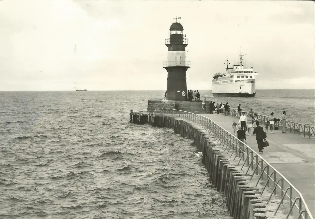 Menschen spazieren auf einem Mole-Strand mit einem Leuchtturm und einem Schiff im Hintergrund.