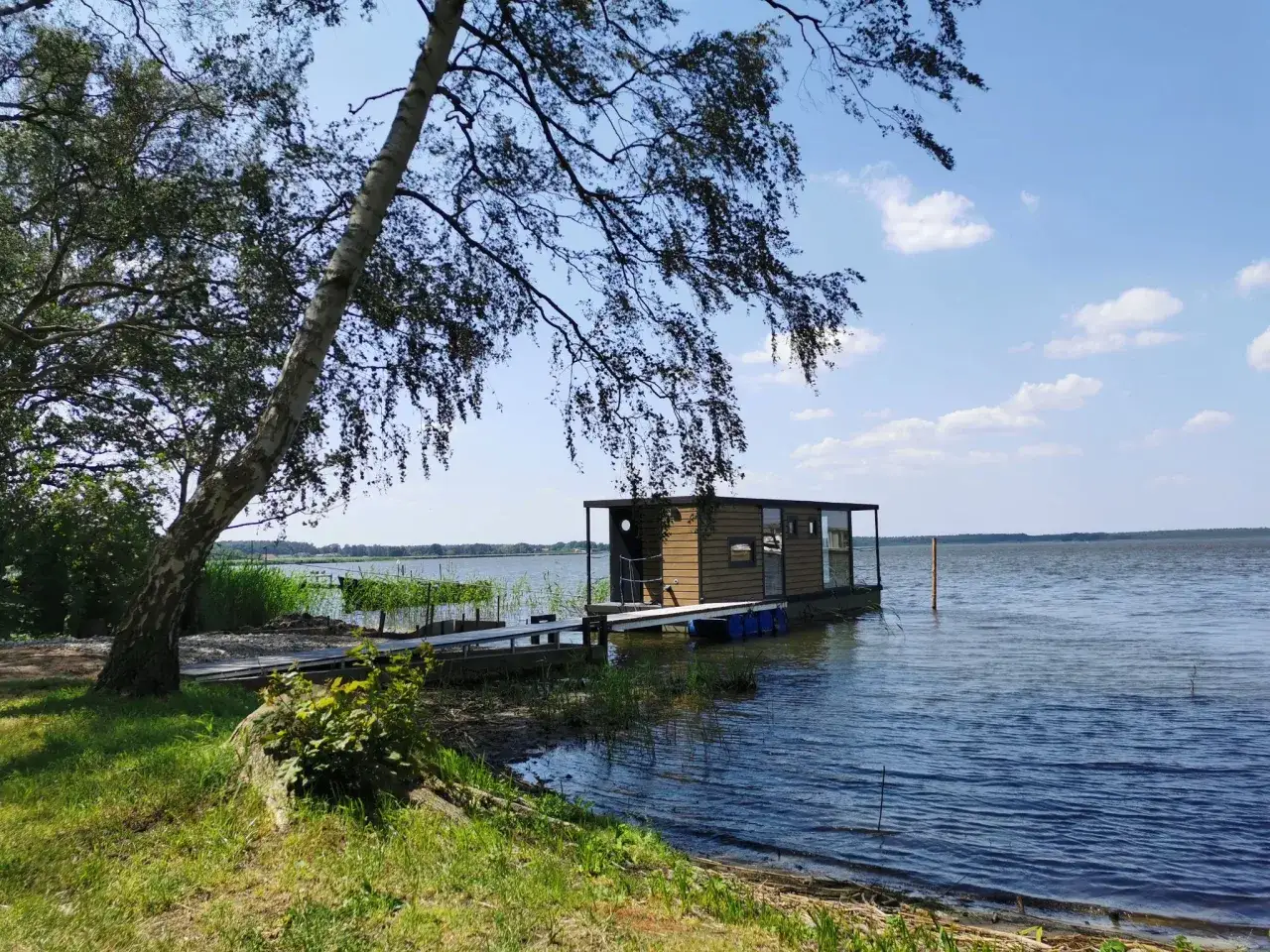 Ein kleines Hausboot liegt am Ufer eines Sees, daneben ein Steg. Ein Baum wirft Schatten auf den grambin strand.