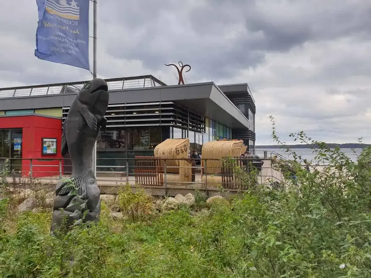 Fischstatue vor modernem Geb&auml;ude am Wasser. Gute Ausflugsziele Ostsee bei schlechtem Wetter, mit Strandk&ouml;rben und Blick aufs Meer.