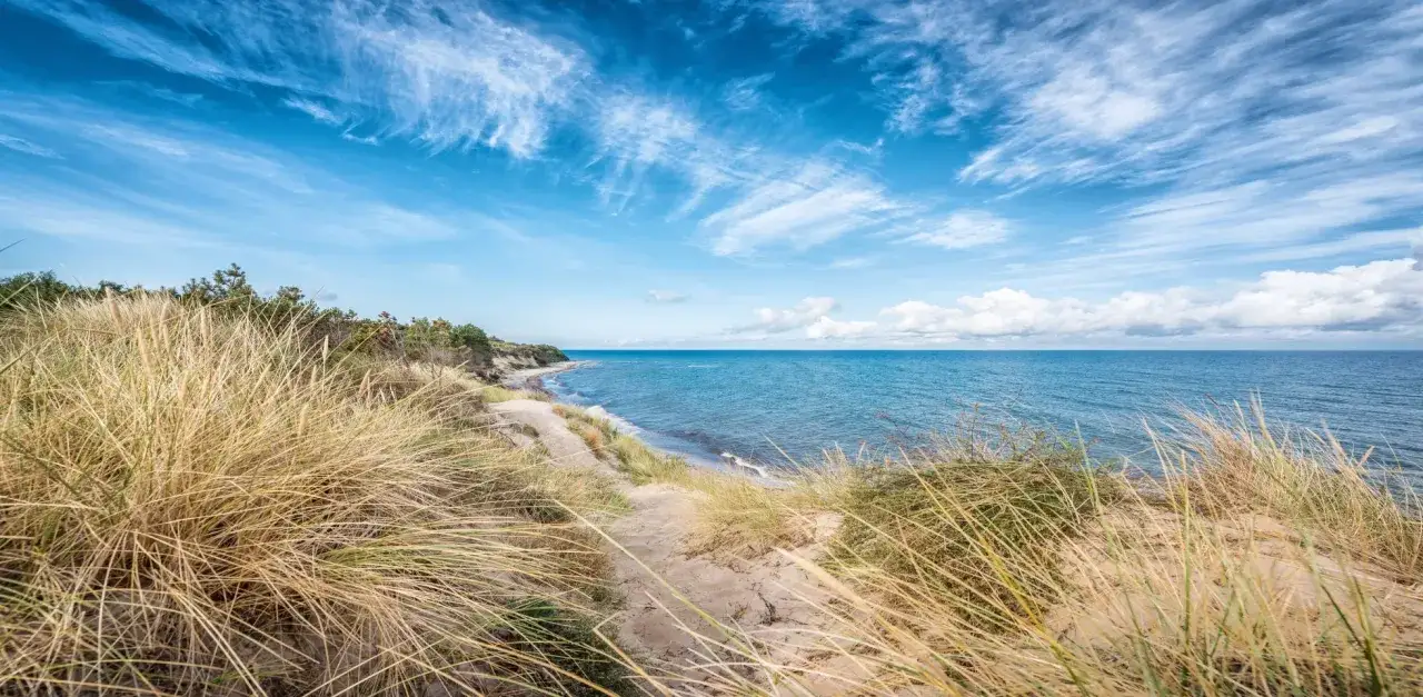 Ein sandiger Pfad f&uuml;hrt durch hohe Gr&auml;ser zum dranske strand. Das Meer glitzert unter einem blauen Himmel mit wei&szlig;en Wolken.