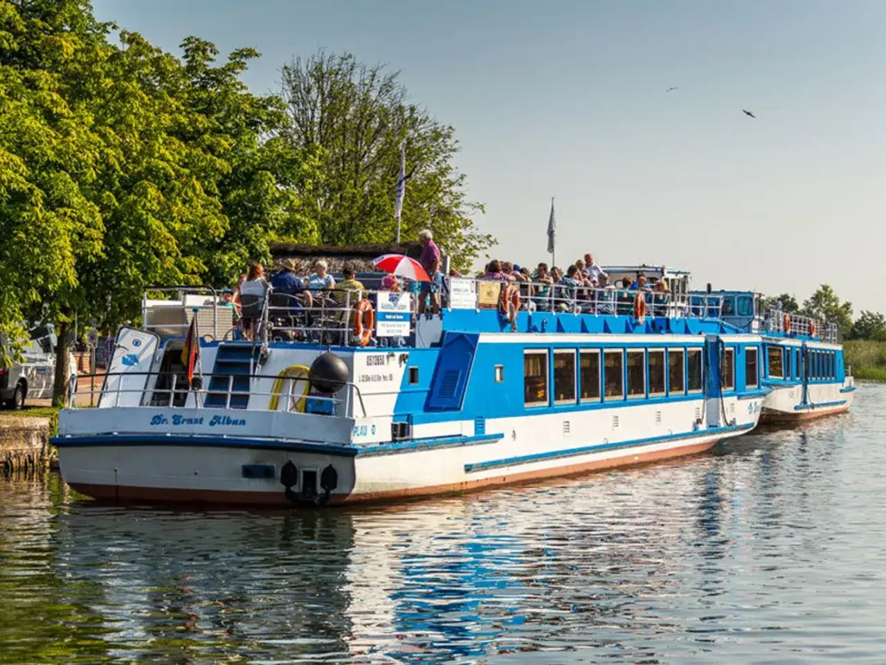 Fahrgastschifffahrt Wichmann: Zwei blau-weiße Schiffe mit Passagieren auf dem Wasser, umgeben von grünen Bäumen und klarem Himmel.
