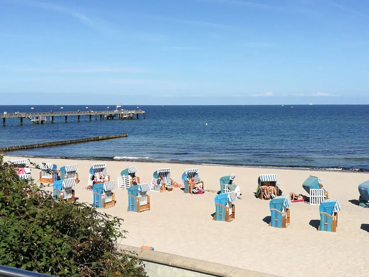Wittenbeck Strand: Strandk&ouml;rbe stehen bereit f&uuml;r sonnige Tage am Meer. Ein langer Pier ragt ins blaue Wasser.