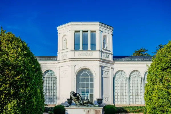 Die Orangerie Putbus, ein strahlend weißes Gebäude mit Skulpturen und großen Fenstern, wird von üppigem Grün eingerahmt.