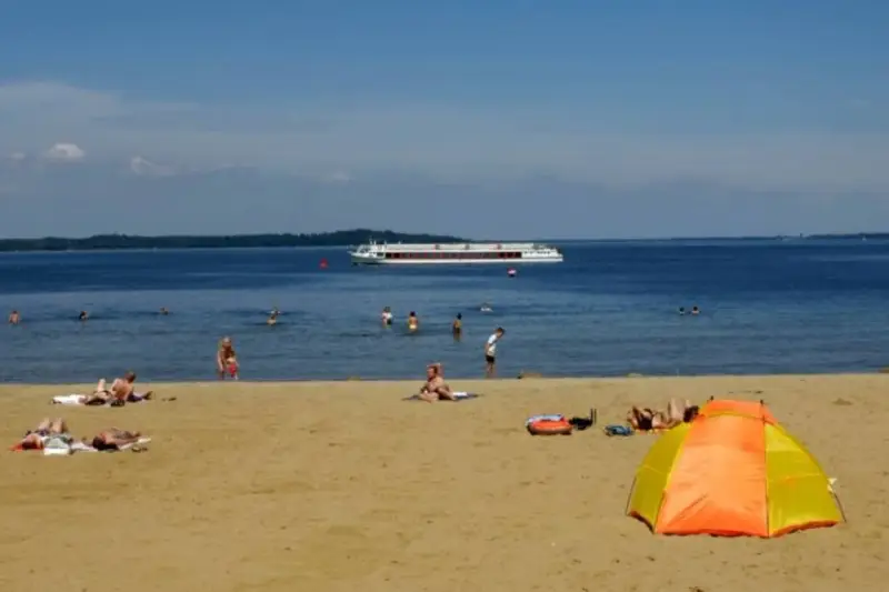Sonne, Sand und ein Schiff auf dem Wasser – ein entspannter Tag am Schweriner Strand mit Badegästen und einer bunten Strandmuschel.