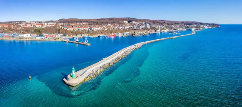 Leuchtturm am Ende einer Mole, die in das klare Wasser vor Sassnitz führt. Boote und die Stadt im Hintergrund.