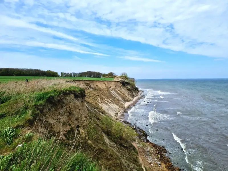 Steilküste mit grasbewachsenem Abhang, der zum steinbeck Strand und dem Meer abfällt. Wolken ziehen über den blauen Himmel.