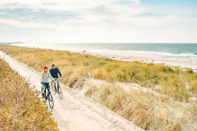 Ein Paar genießt seine baltischen Abenteuer auf Fahrrädern entlang eines sandigen Weges am Meer, umgeben von Dünen und Gräsern.